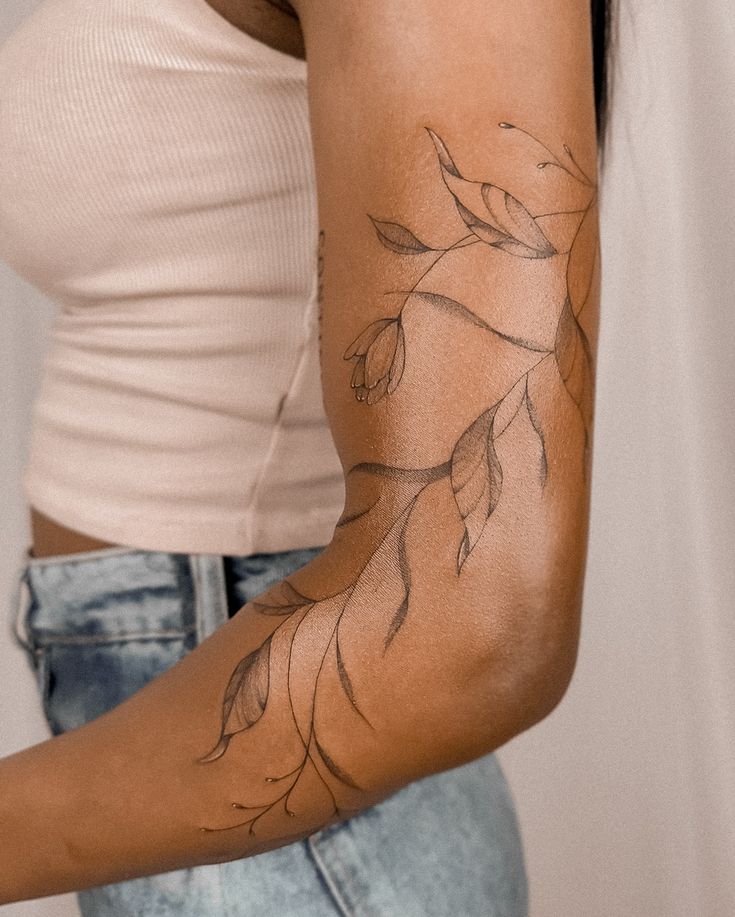 Elegant botanical tattoo with leaves on a woman's upper arm, wearing a beige top and jeans.
