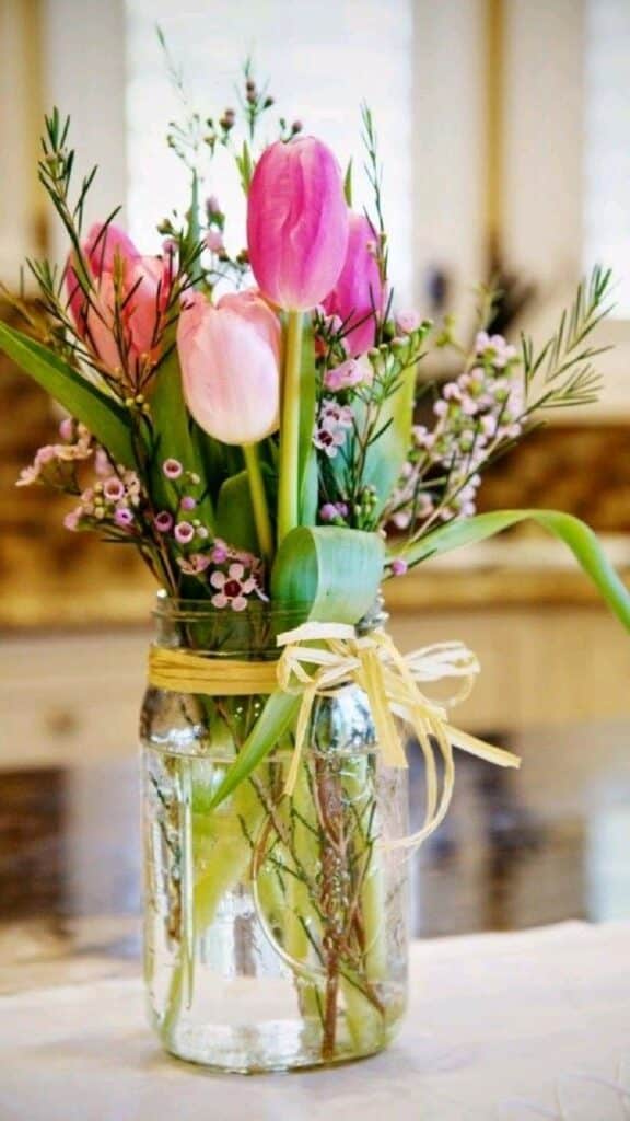 Pink tulips and greenery in a glass jar vase with a raffia bow on a kitchen counter.