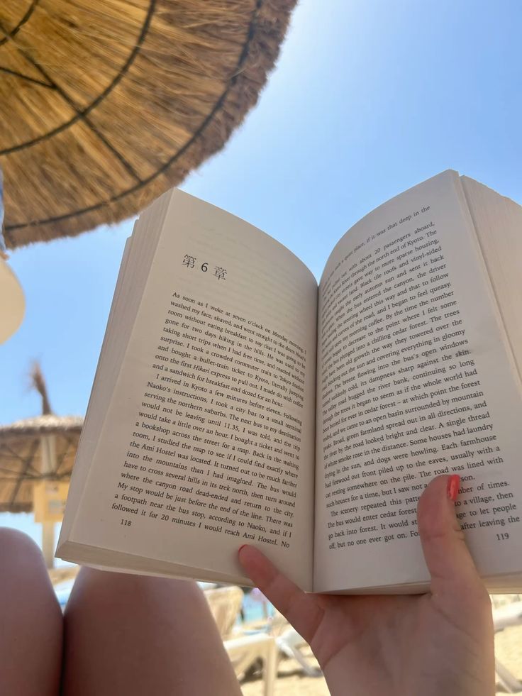 Reading a book on a sunny beach under a thatched umbrella, with a clear blue sky above.