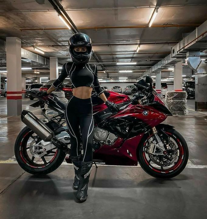 Motorcyclist in black gear stands beside a sleek red and black sports bike in a parking garage.