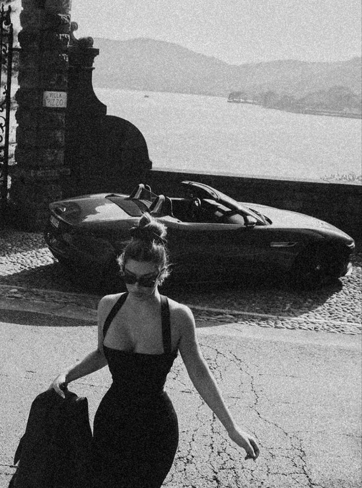 Woman in stylish dress walking near convertible car by scenic lakeside.
