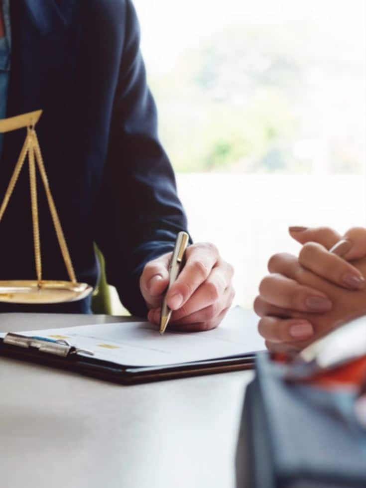 Lawyer meeting with client, holding scales and pen, discussing legal documents on clipboard.