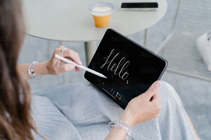 Person using a stylus to write Hello on a tablet screen, with a drink on a table nearby.