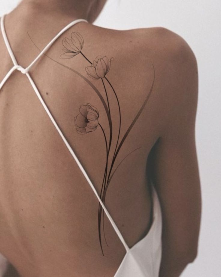 Elegant floral tattoo on back with white strappy top, showcasing intricate flower design and delicate lines.