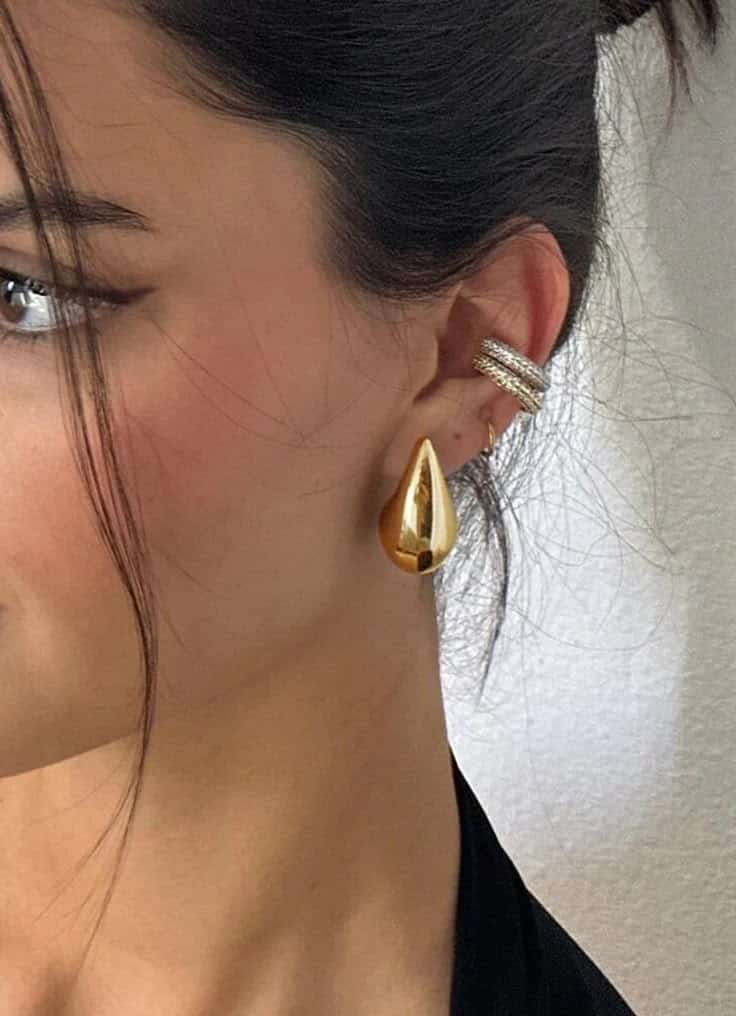 Woman wearing stylish gold drop earring and multiple silver ear cuffs on a white background.