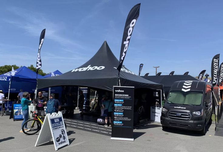 Outdoor cycling event with Wahoo tent, display boards, and visitors engaging. Sunny day with clear blue sky.