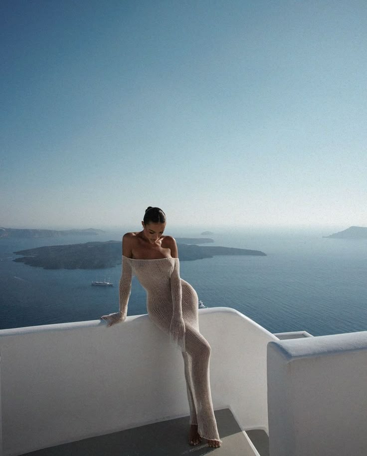 Woman in elegant dress on balcony overlooking serene seascape, surrounded by clear blue skies and distant islands.