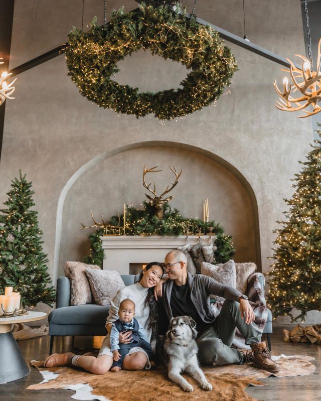 Family and dog by the fireplace in a festive living room with Christmas trees and wreath. Cozy holiday setting.