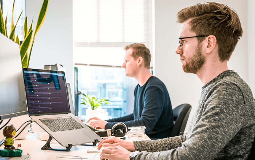 Two men working at computers in a bright, modern office space, focused on coding tasks.