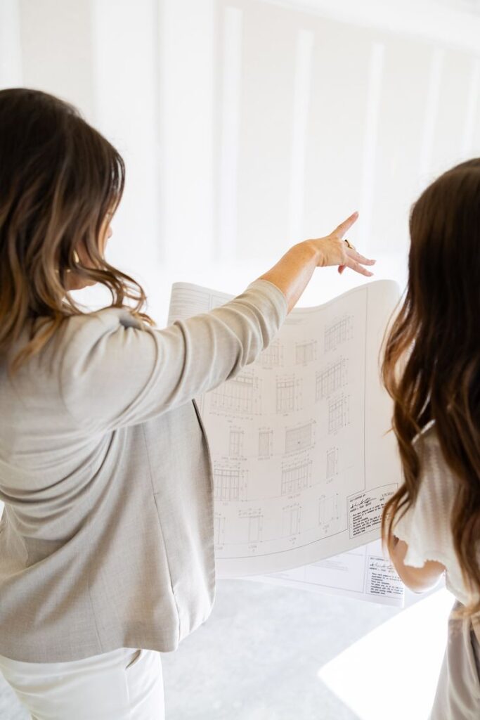 Two women discussing architectural plans indoors, one pointing at details on the blueprint, wearing professional attire.