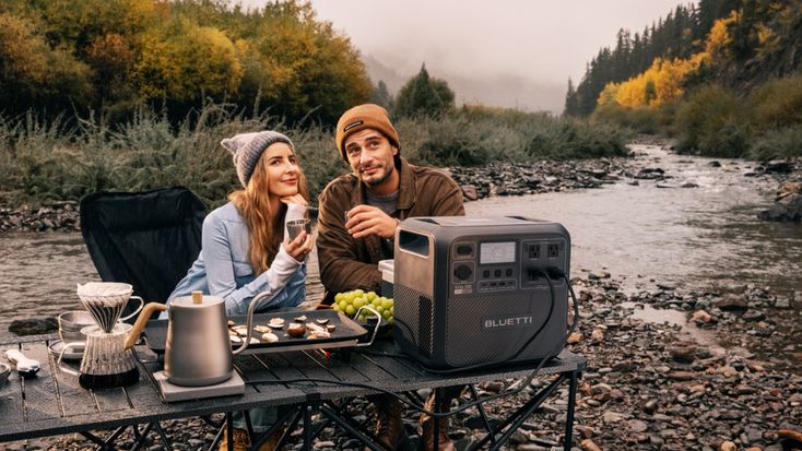 Couple enjoying a riverside picnic with a portable power station and snacks on a table, surrounded by autumn foliage.