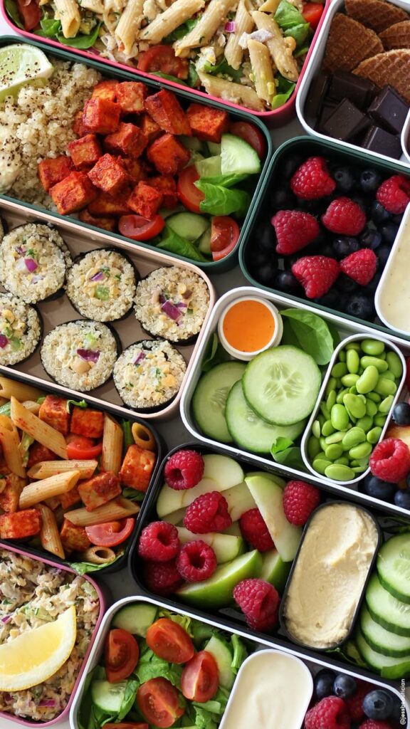 Colorful vegan lunch boxes with pasta, fruits, salads, quinoa rolls, and dipping sauces for a healthy meal prep.
