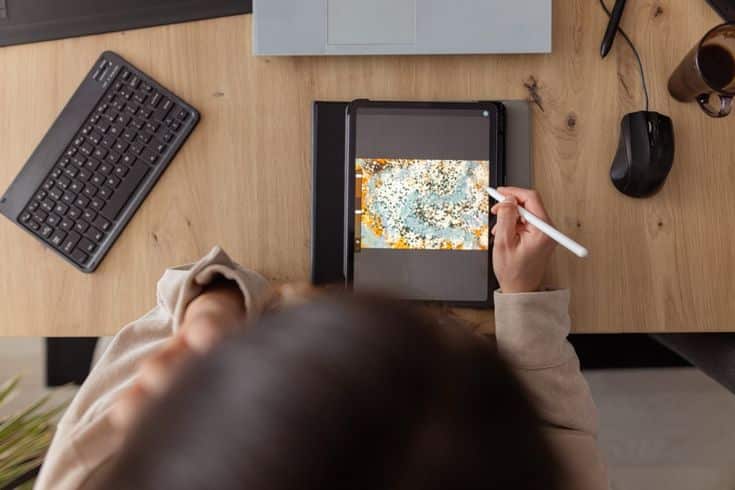 Person using a stylus on a digital tablet with colorful design on screen, with keyboard and mug nearby on a wooden desk.