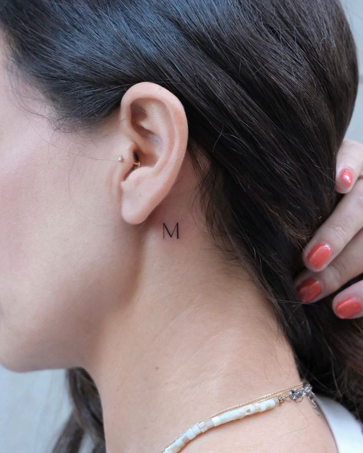 Close-up of a minimalist M tattoo behind a woman's ear with a small ear piercing and painted nails.