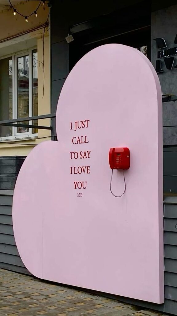 Pink heart-shaped wall with red phone and I just call to say I love you text; romantic outdoor installation.