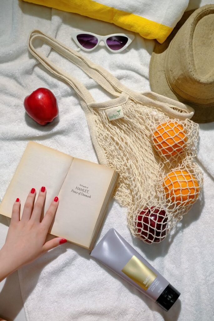 Summer essentials: open book, mesh bag with fruits, sunglasses, hat, and sunscreen on a white towel.