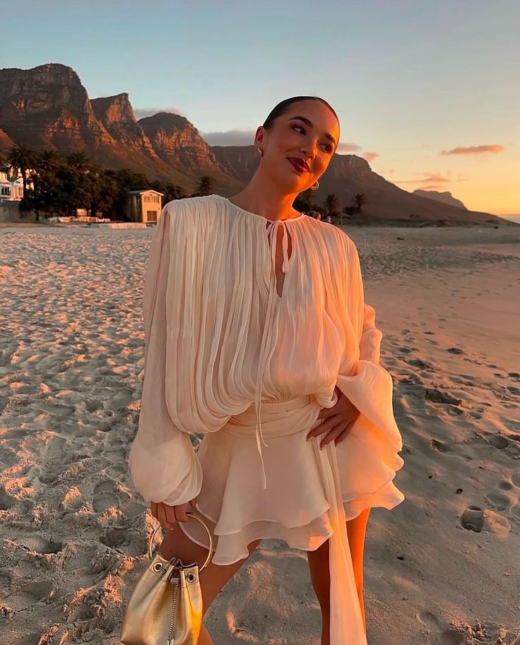 Woman in white dress on a beach at sunset, with mountains in the background.
