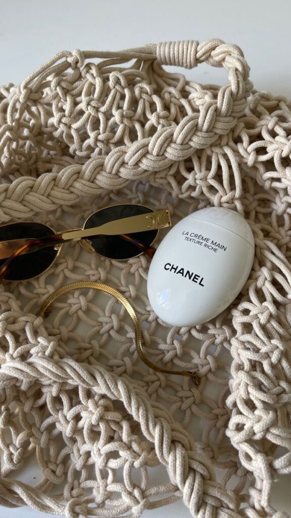 Chic summer essentials: macramé bag, sunglasses, gold bracelet, and Chanel hand cream on a white background.