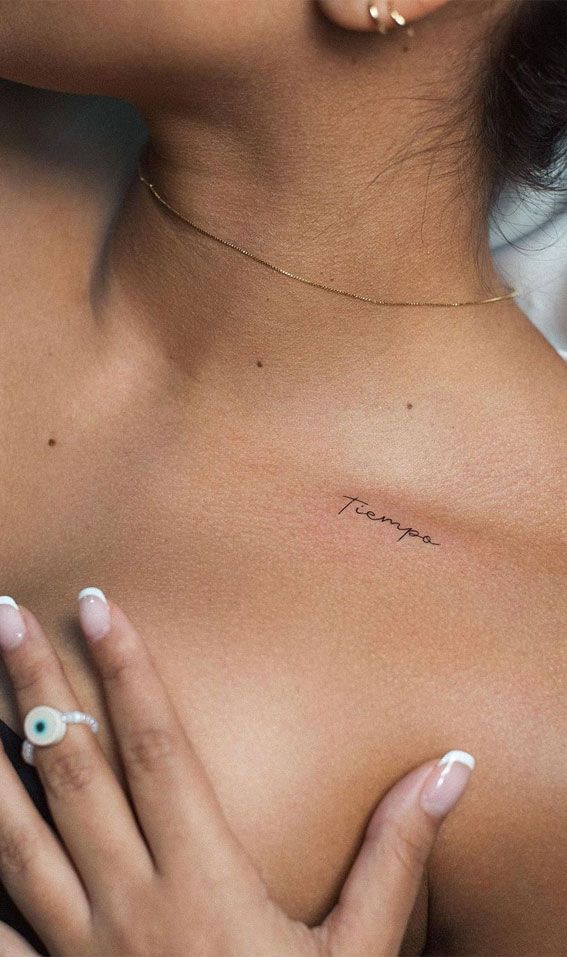 Elegant collarbone tattoo with Tiempo script, delicate gold necklace, and manicured hand touching skin.