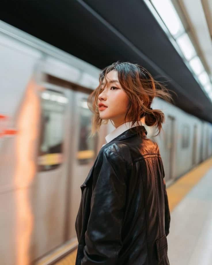 Woman in a black leather jacket standing at a subway platform as a train passes by, urban setting.