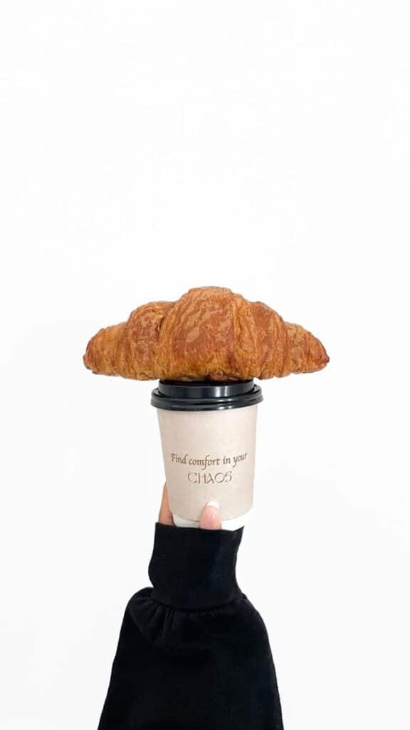 Hand holds coffee cup topped with croissant, text reads Find comfort in your chaos. Minimalist breakfast concept.