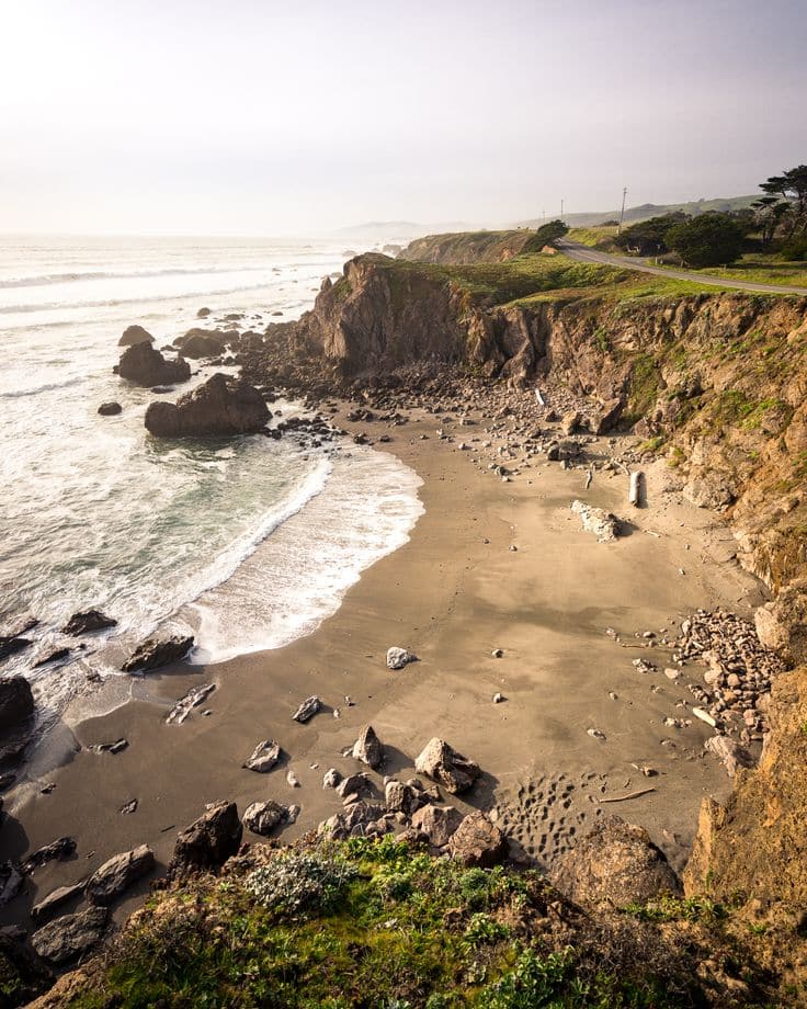 Scenic coastal view with rocky cliffs, sandy beach, and gentle ocean waves under a clear sky.
