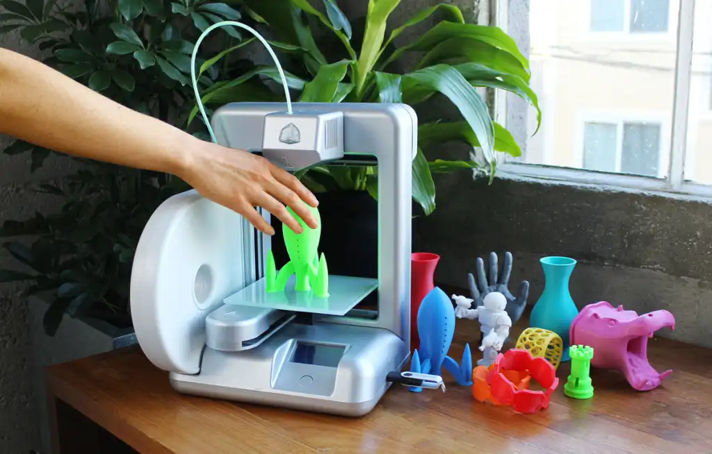 Person removing a green 3D printed rocket from a printer, surrounded by colorful 3D objects on a wooden table.