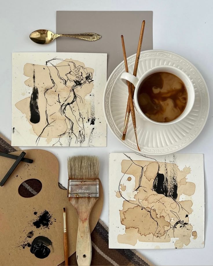 Artistic setup with abstract drawings, brushes, coffee, and gold spoon on white and brown background.
