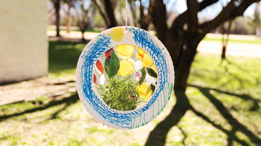 Colorful flower suncatcher hanging outdoors, reflecting sunlight through translucent floral design against tree backdrop.