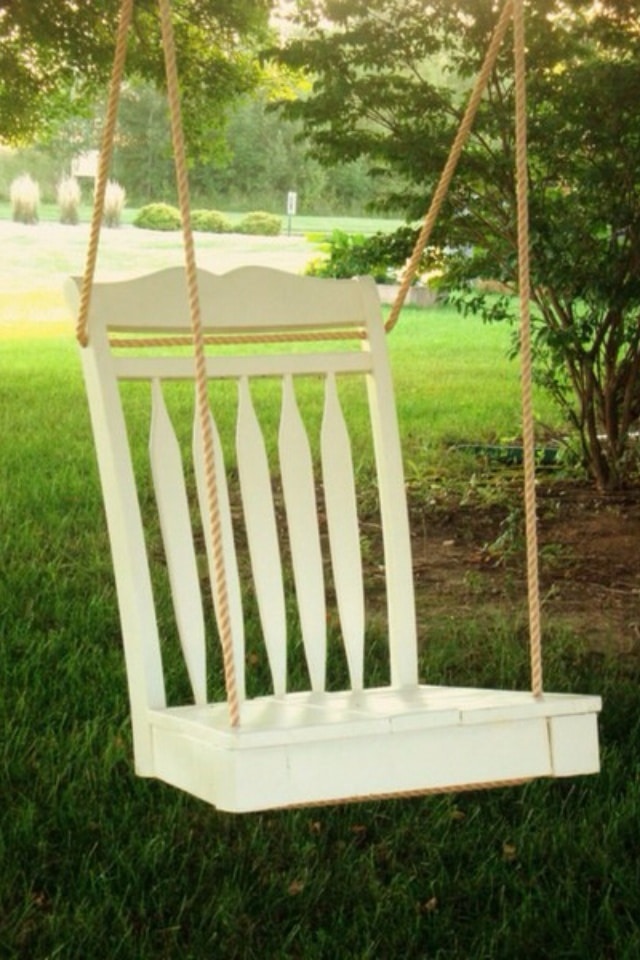 White wooden chair swing hanging in a garden, surrounded by green grass and trees, bathed in sunlight.