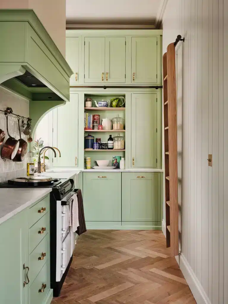 Cozy kitchen with pastel green cabinets, open pantry, and herringbone wood floor for a charming, rustic feel.