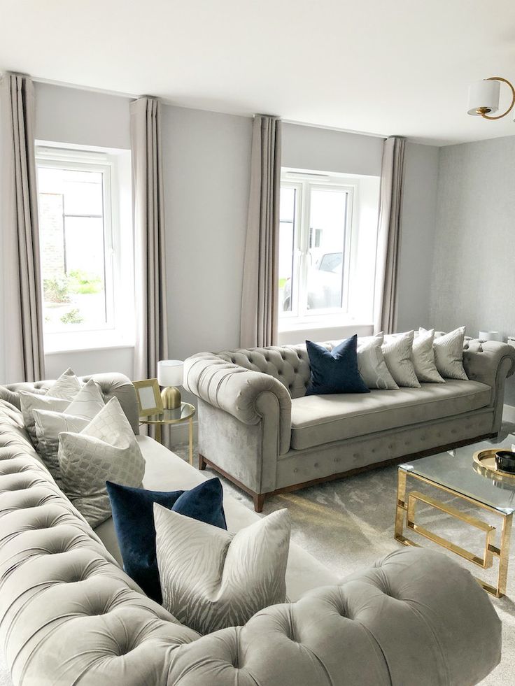 Elegant living room with tufted gray sofas, plush pillows, and glass coffee table near large windows and neutral curtains.