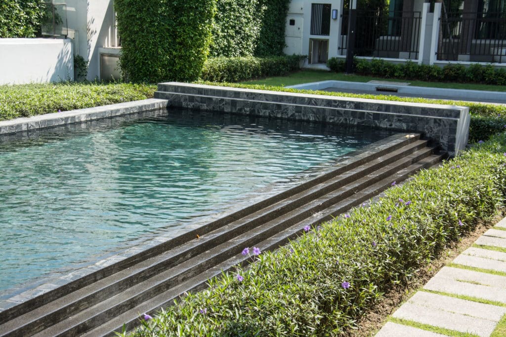 Modern rectangular swimming pool with stone steps, surrounded by lush greenery and purple flowers in a serene backyard.