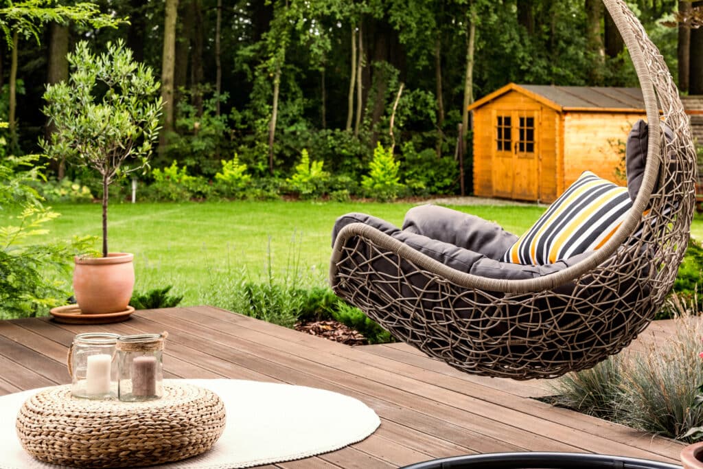 Depositphotos_192576756_XL Cozy backyard with a wicker hanging chair, striped cushion, and a wooden deck, set against a lush green garden backdrop. | Sky Rye Design Cozy backyard with a wicker hanging chair, striped cushion, and a wooden deck, set against a lush green garden backdrop.