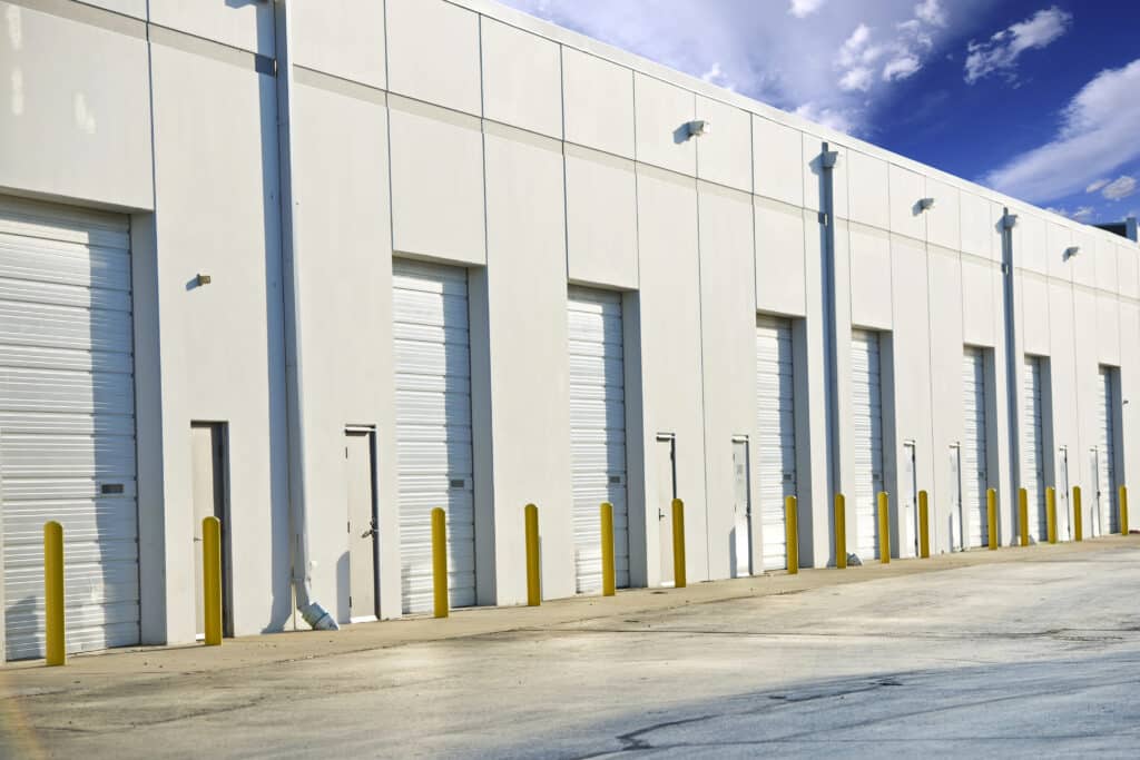 Large industrial warehouse exterior with multiple closed loading doors and yellow bollards under a clear blue sky.
