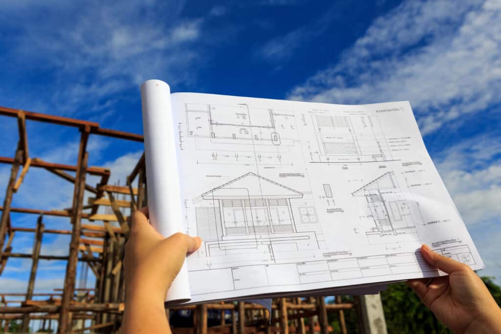 Depositphotos_32226229_XL Architectural blueprint held in front of a house under construction, set against a blue sky background. | Sky Rye Design Architectural blueprint held in front of a house under construction, set against a blue sky background.