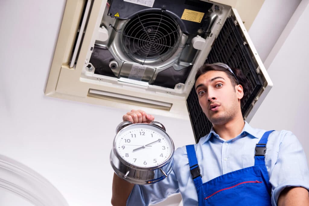 Technician holding clock beneath opened ceiling AC unit, emphasizing timely HVAC maintenance and repair services.