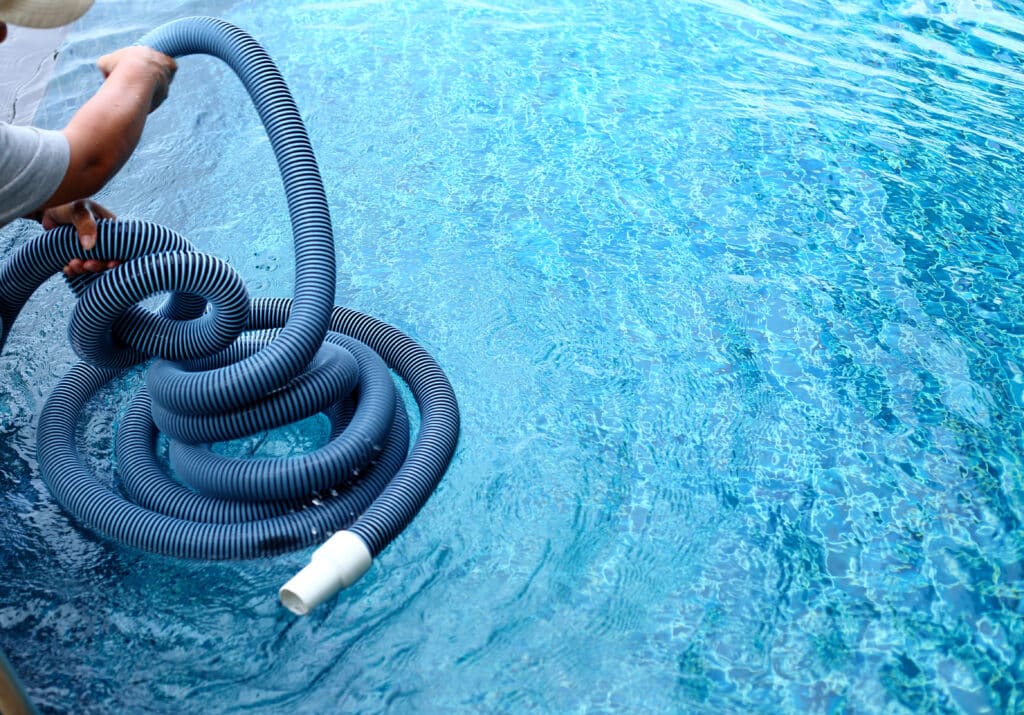 Person cleaning a swimming pool with a flexible hose, clear blue water reflecting sunlight.