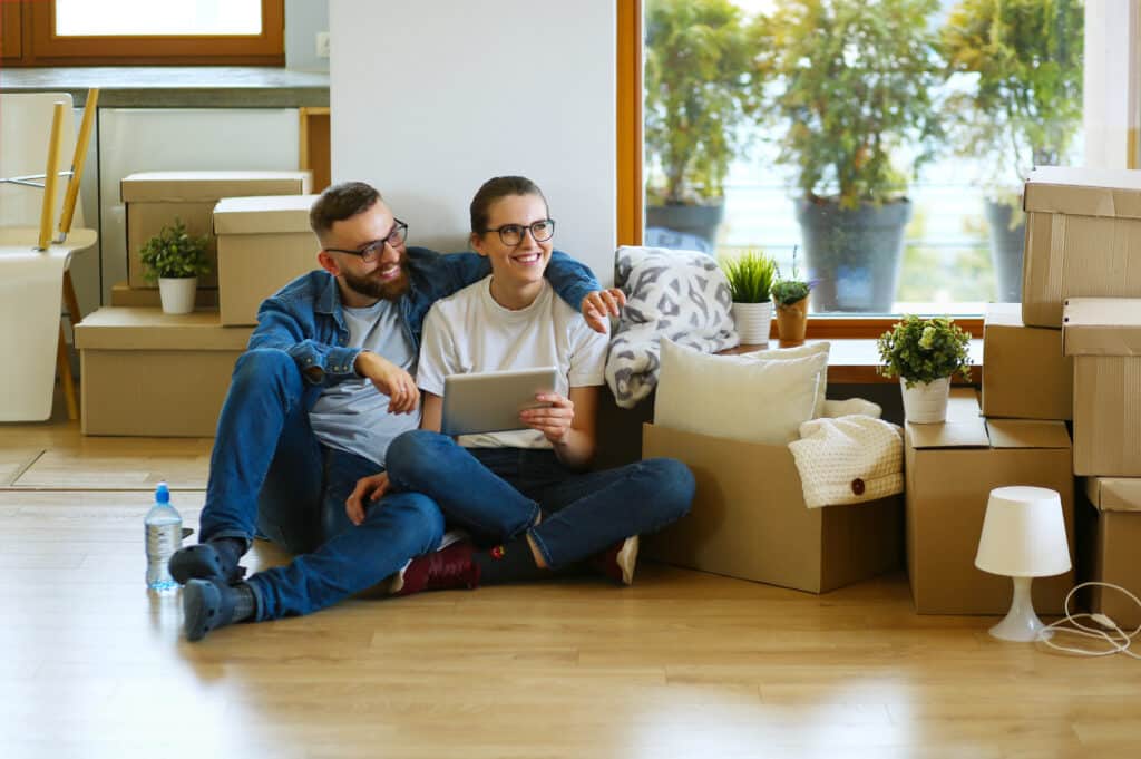 Happy couple sitting among moving boxes, using a tablet and smiling in their new home.
