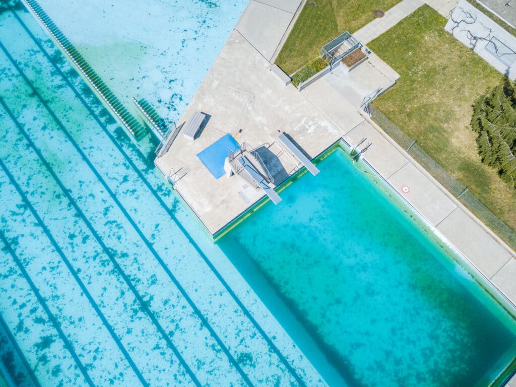 Depositphotos_367519510_XL Aerial view of a swimming pool with diving boards surrounded by grass and walkways. | Sky Rye Design Aerial view of a swimming pool with diving boards surrounded by grass and walkways.