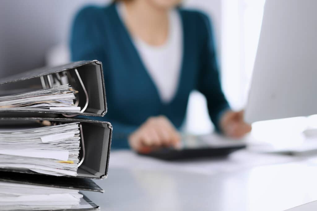 Depositphotos_390036090_XL Stacked binders and a person working at a desk, representing office organization and productivity. | Sky Rye Design Stacked binders and a person working at a desk, representing office organization and productivity.