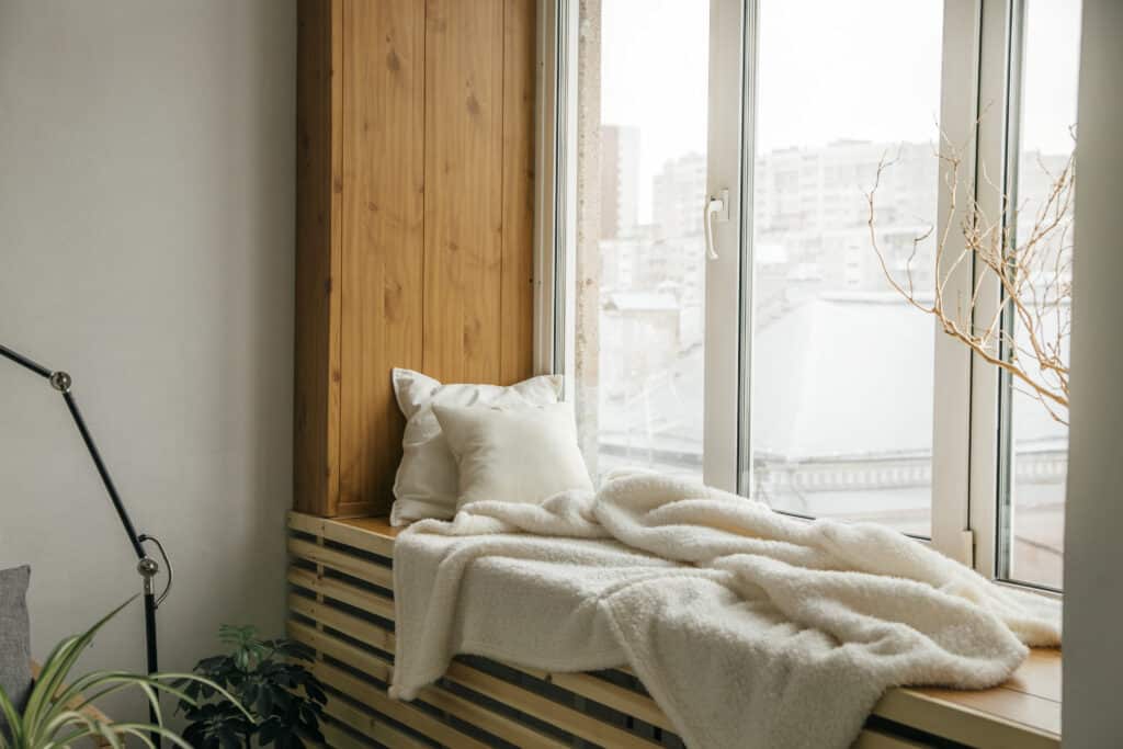 Cozy window seat with white pillows and blanket, overlooks cityscape through large window, creating a peaceful reading nook.