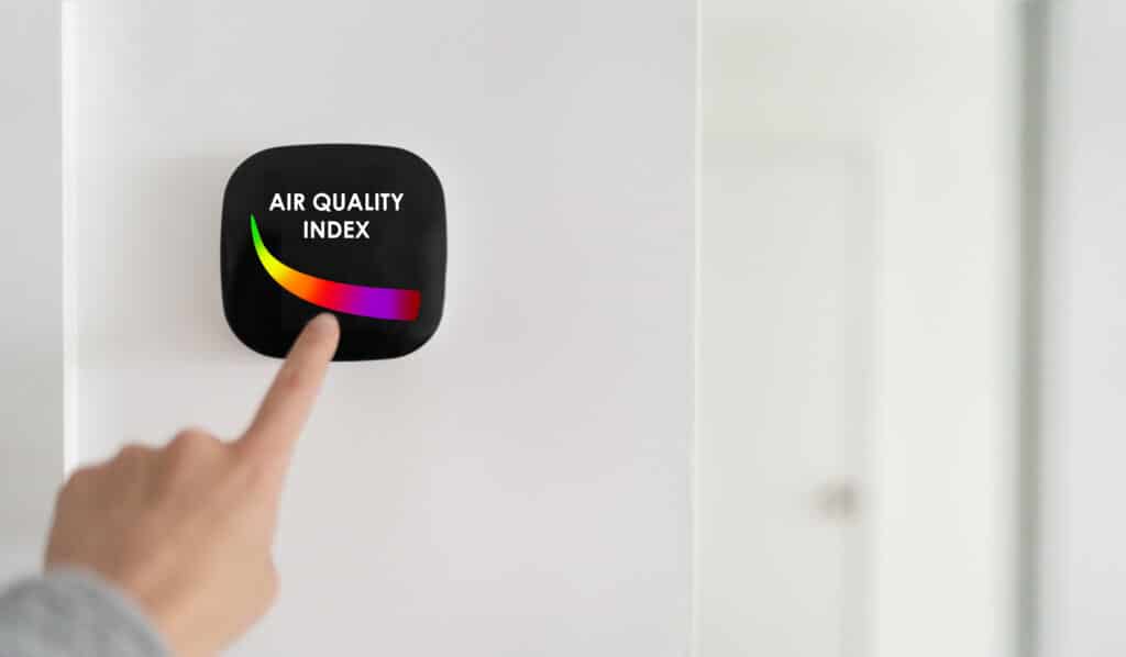Finger touching air quality index monitor with a colorful gauge on white wall.