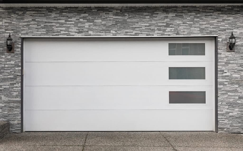Modern white garage door with three small windows, set in a stone wall exterior.