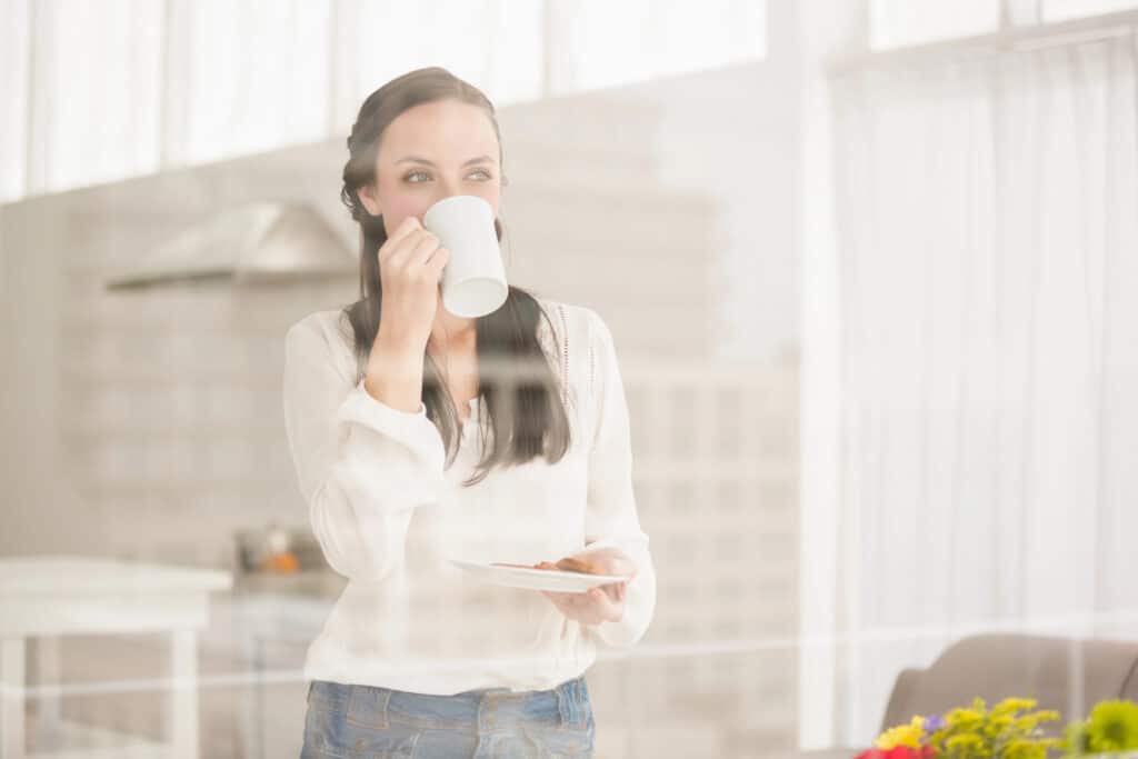 Woman enjoying a hot drink by a window with a small plate in hand. Bright and cozy indoor setting.