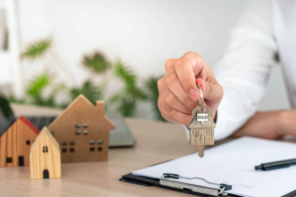 Hand holding house key with 'Sold' label, miniature houses on desk; concept of real estate transaction and property sale.