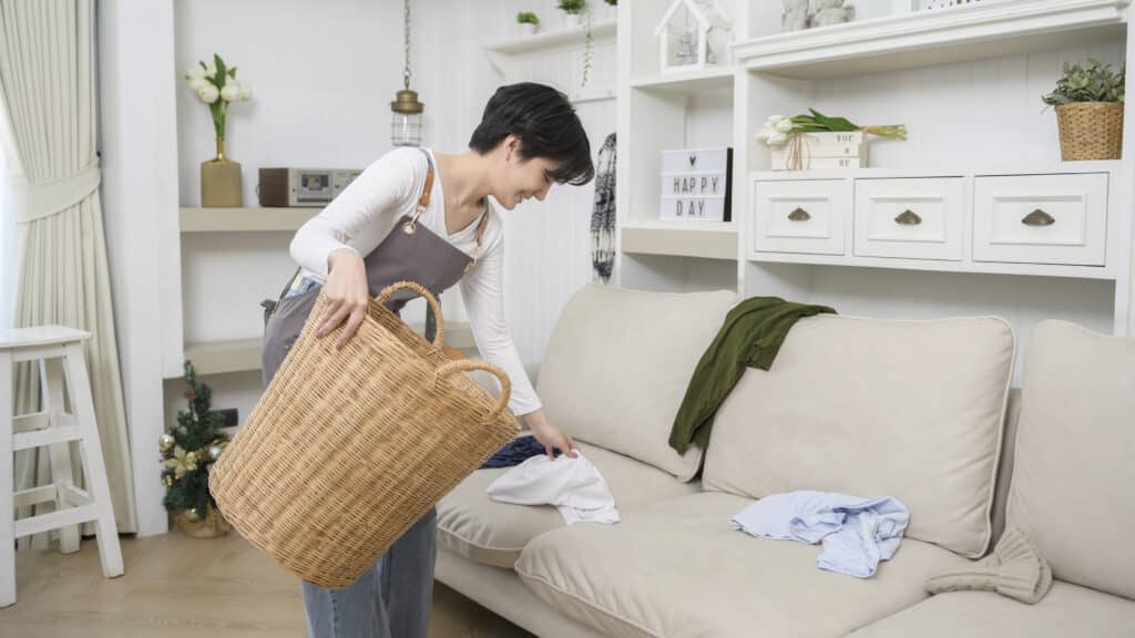 Woman tidying a cozy living room, organizing clothes into a wicker laundry basket on a beige couch.