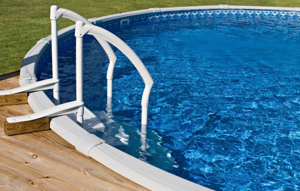 Above ground swimming pool with wooden deck and white ladder, surrounded by green grass.