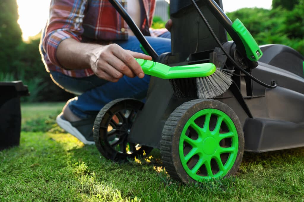 Person cleaning a lawn mower with a brush on grass, focusing on maintenance.