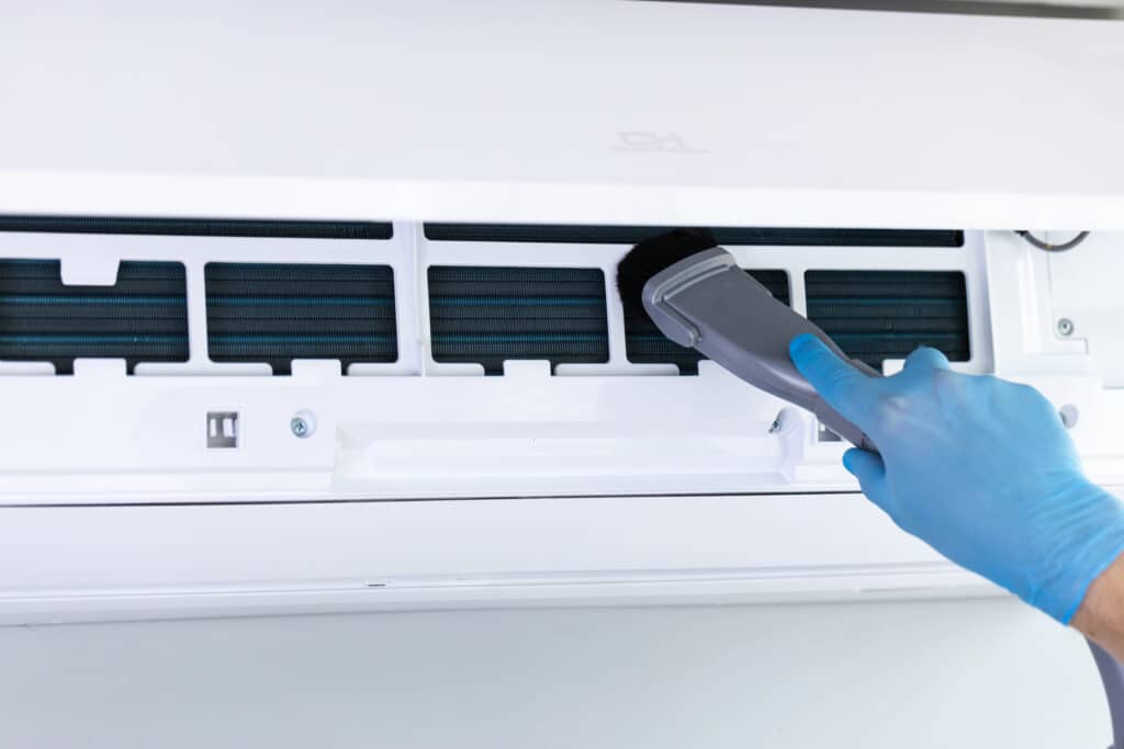 Person cleaning air conditioner filter with brush wearing gloves.